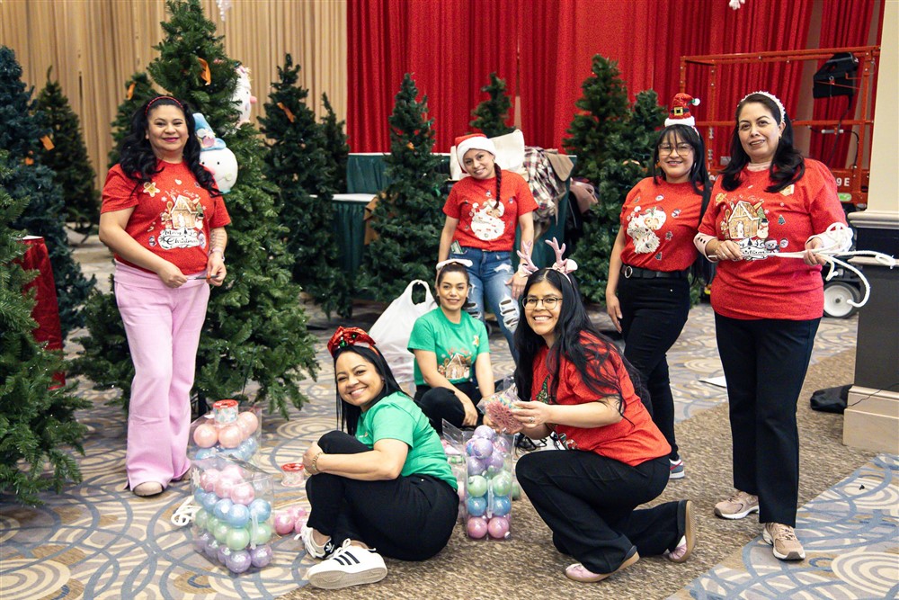 A group of Operation North Pole volunteers in matching festive shirts smiling together while decorating with ornaments and Christmas trees